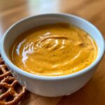 Close-up of rich, orange beer cheese dip served in a white bowl next to salted pretzels.