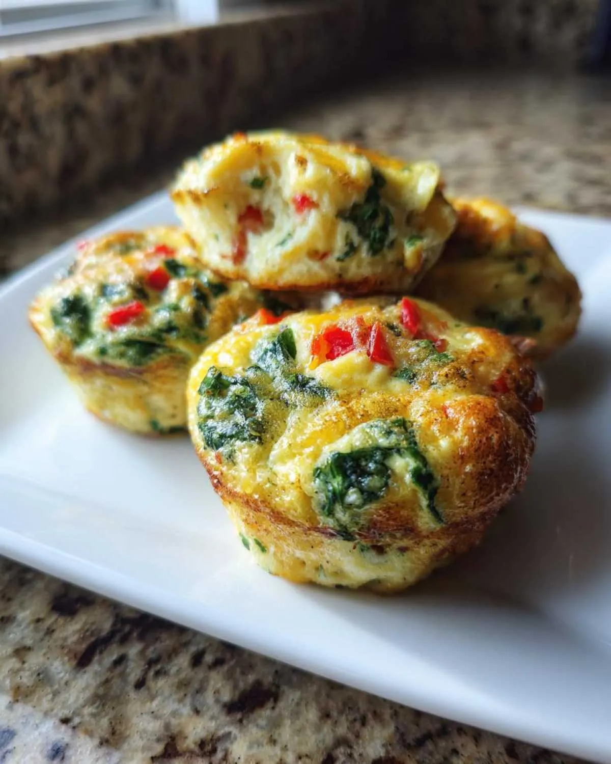 Four baked egg muffin cups filled with spinach and red peppers, stacked on a white plate.