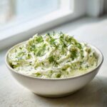 A bowl of creamy, light green pickle dip generously topped with fresh dill sprigs.