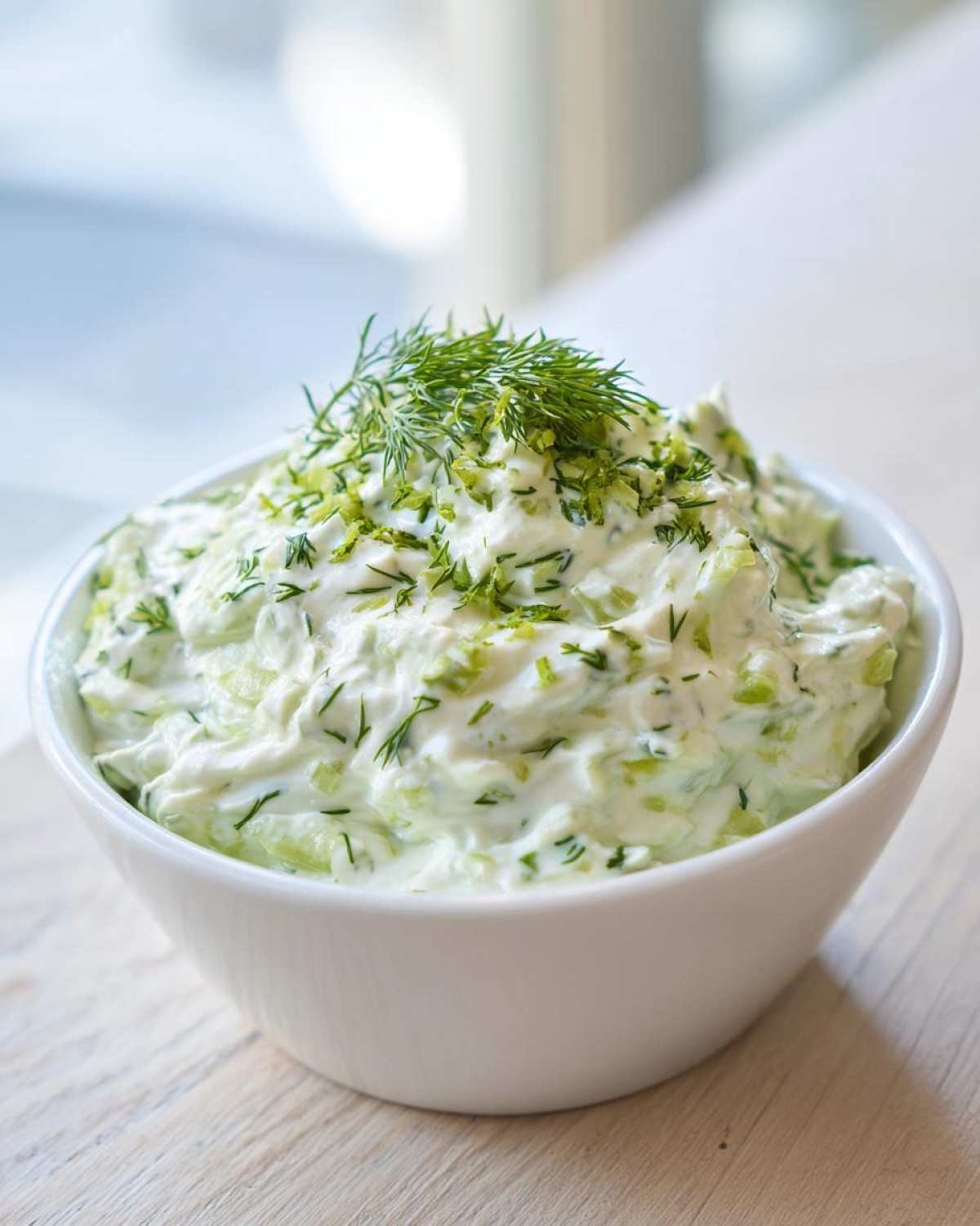 Close-up of creamy, chunky pickle dip topped with fresh dill in a white bowl.