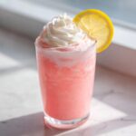 A tall glass of frosty pink lemonade topped with whipped cream and a lemon slice.