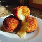 Close-up of two golden-brown, deep-fried rice balls, one cut open revealing melted cheese oozing out.