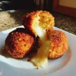 Close-up of two golden-brown, deep-fried rice balls, one cut open revealing melted cheese oozing out.
