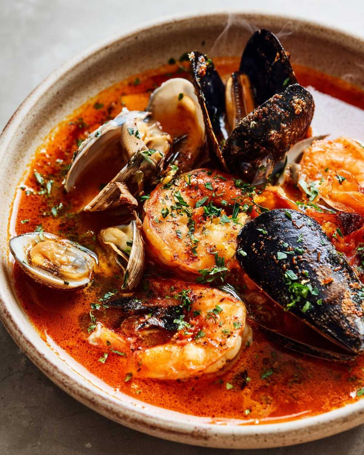 Close-up of a steaming bowl of rich, red seafood soup featuring large shrimp, mussels, and clams, garnished with parsley.