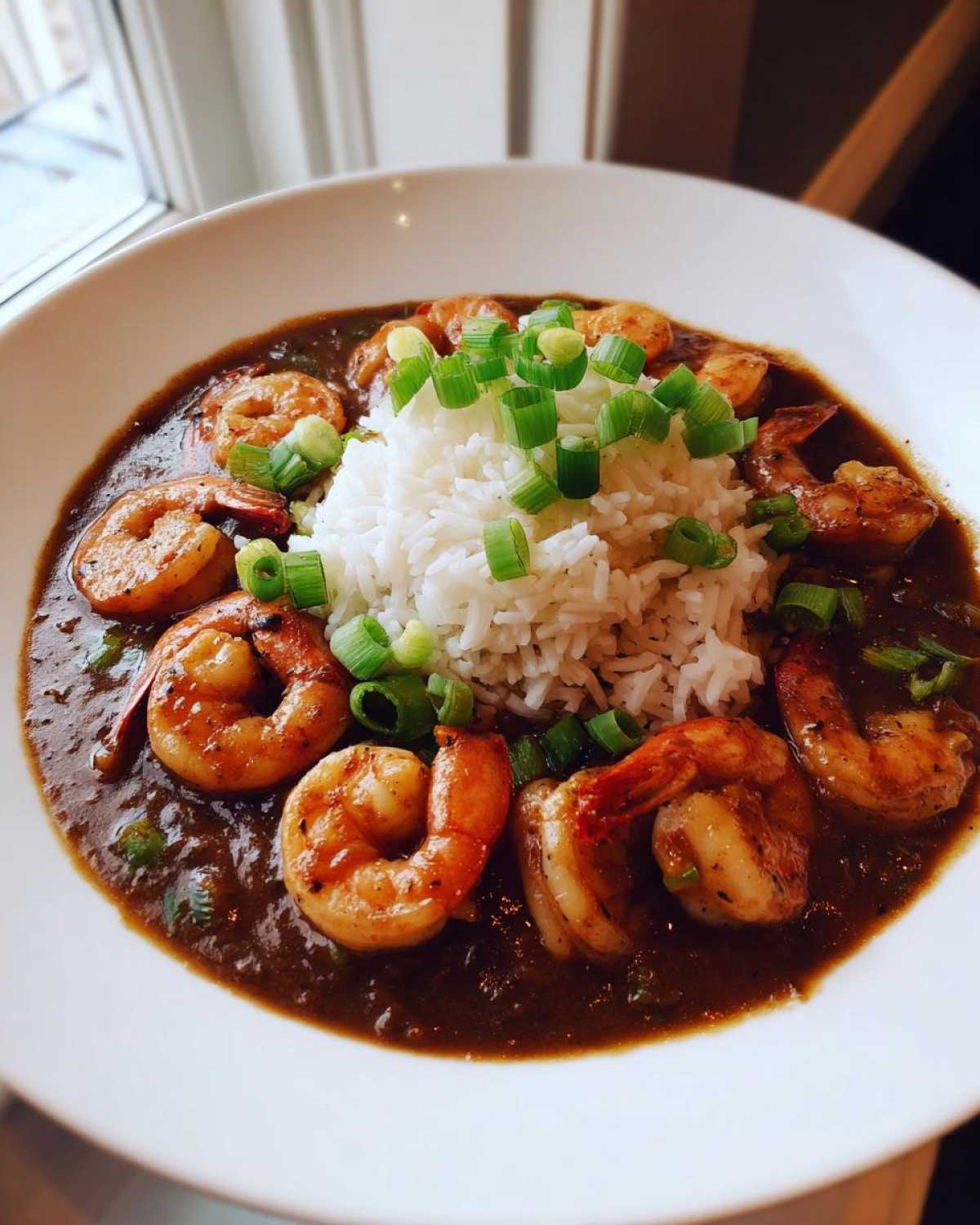 A close-up of a bowl of rich, dark shrimp etouffee served with a mound of white rice and topped with sliced green onions.
