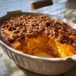 A scoop removed from a baked sweet potato souffle topped with crunchy pecans in a white oval dish.
