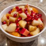 Close-up of a white bowl filled with amazing winter fruit salad featuring diced pears, pomegranate seeds, and orange segments.