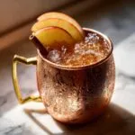 Close-up of a frosty copper mug filled with an apple cider moscow mule, garnished with apple slices and a cinnamon stick.