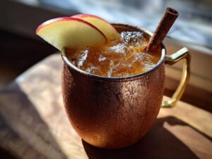 Close-up of a frosty copper mug filled with an apple cider moscow mule, garnished with apple slices and a cinnamon stick.