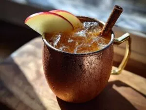 Close-up of a frosty copper mug filled with an apple cider moscow mule, garnished with apple slices and a cinnamon stick.