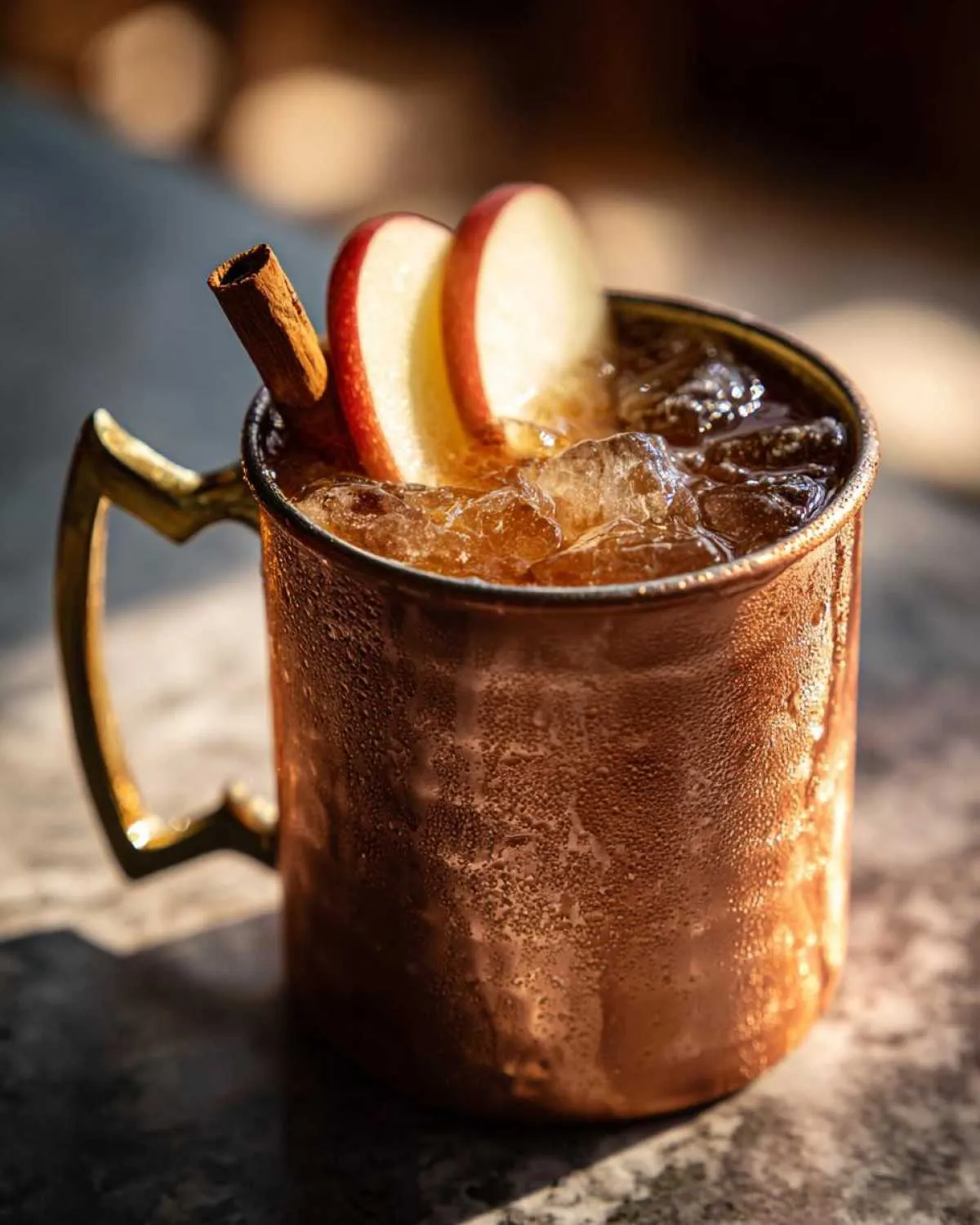 Close-up of a frosty copper mug filled with an apple cider moscow mule, garnished with apple slices and a cinnamon stick.
