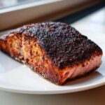 A thick fillet of blackened salmon resting on a white plate, showing a dark, spicy crust and moist pink interior.