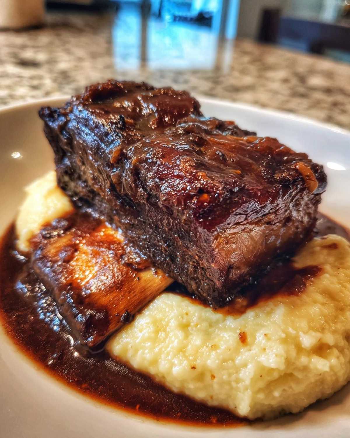 A single, thick braised short ribs piece covered in dark glaze resting on creamy polenta with rich sauce.