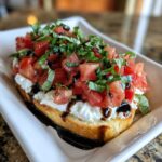 A single piece of toasted bread topped with creamy cheese, diced tomatoes, fresh basil, and balsamic glaze, representing a bruschetta dip serving.