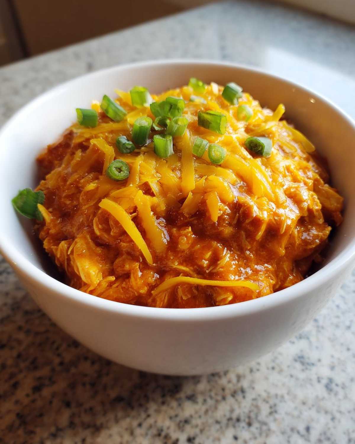 Close-up of a bowl of rich, orange buffalo chicken chili topped with shredded cheddar cheese and green onions.