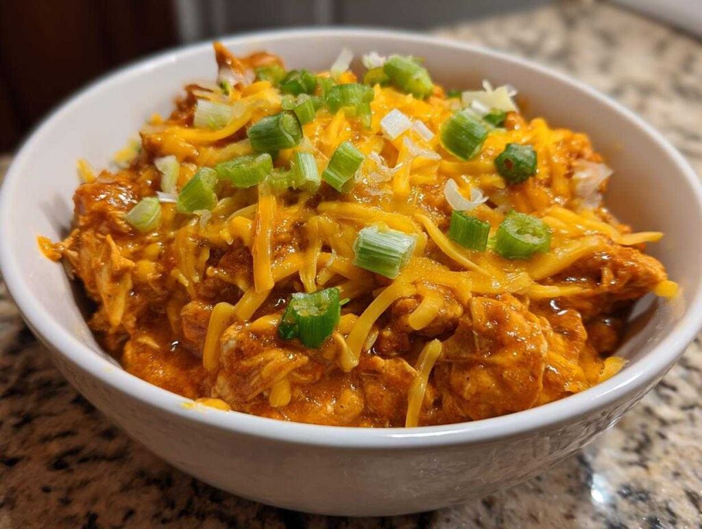 Close-up of a white bowl filled with rich, orange buffalo chicken chili topped with melted shredded cheddar cheese and chopped green onions.