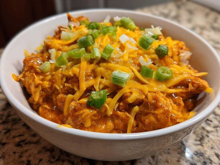 Close-up of a white bowl filled with rich, orange buffalo chicken chili topped with melted shredded cheddar cheese and chopped green onions.