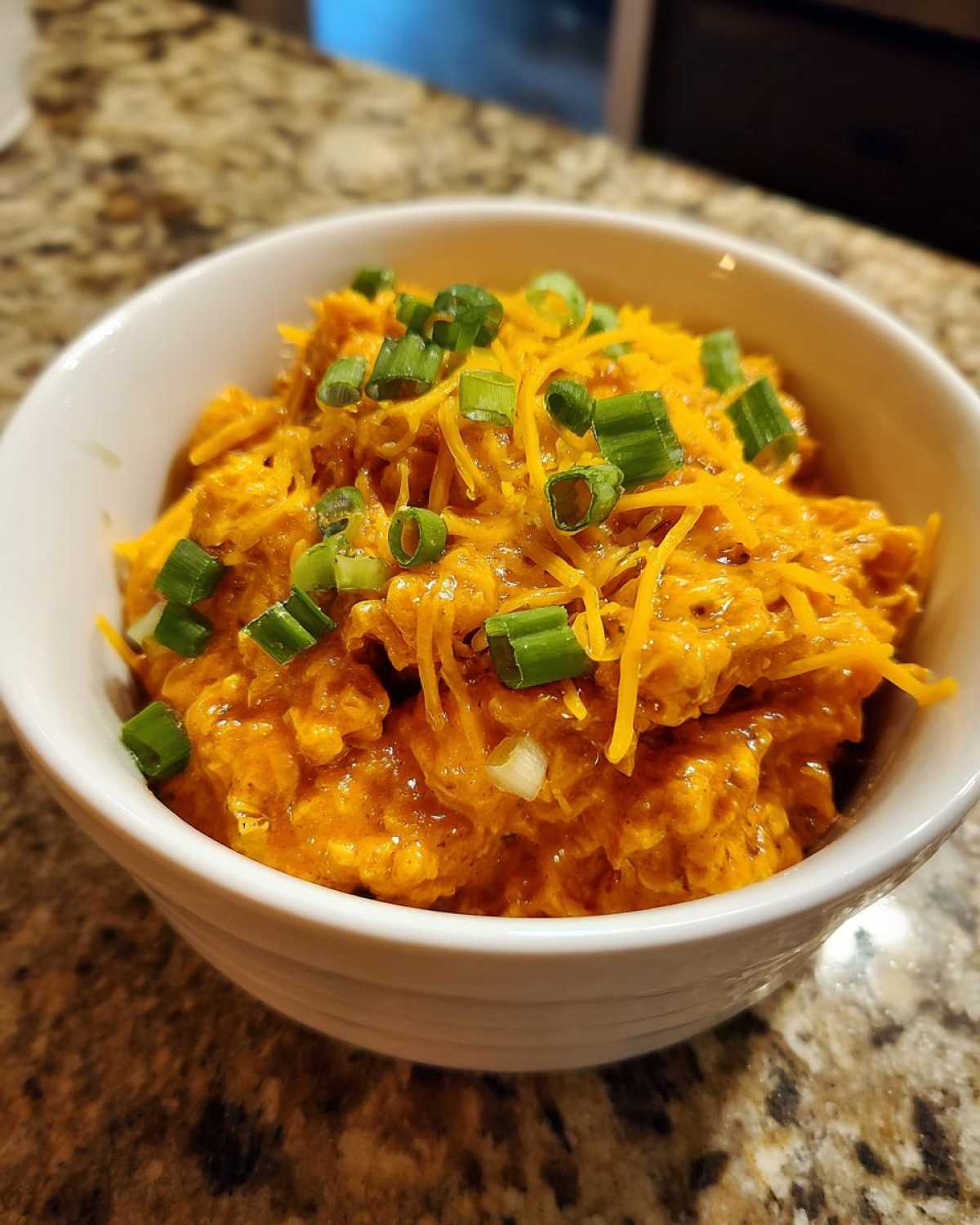 A close-up of a white bowl filled with creamy buffalo chicken chili, topped with shredded cheddar cheese and chopped green onions.