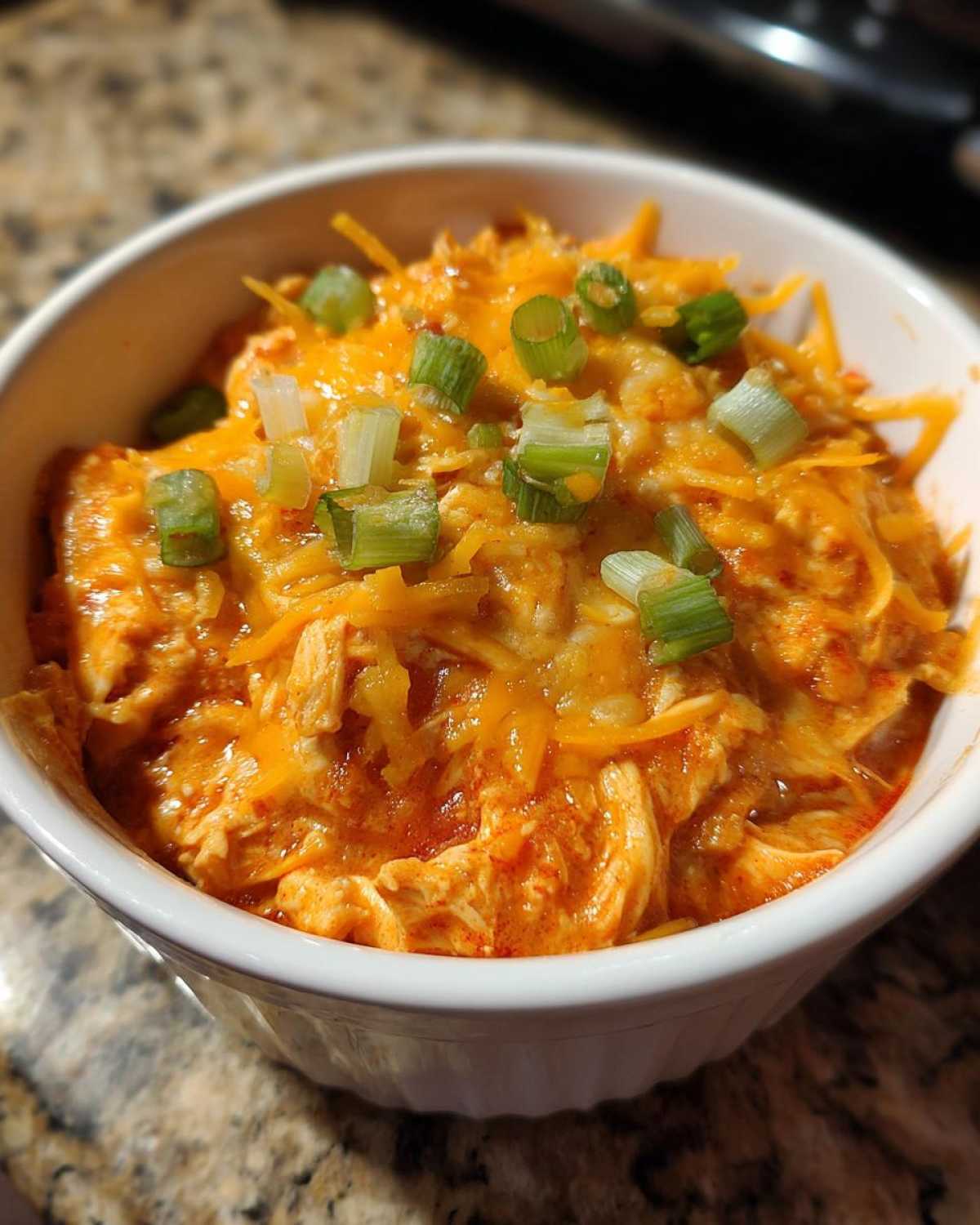 Close-up of a white bowl filled with rich orange buffalo chicken chili, topped with melted cheddar cheese and sliced green onions.