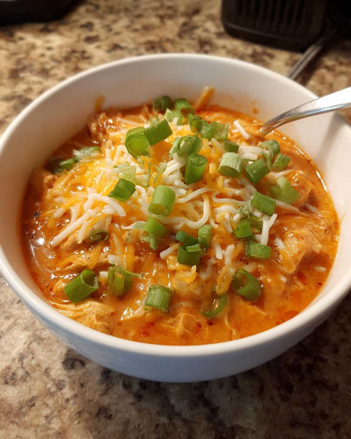 A close-up of a white bowl filled with creamy, orange buffalo chicken chili, topped with shredded cheese and fresh green onions.