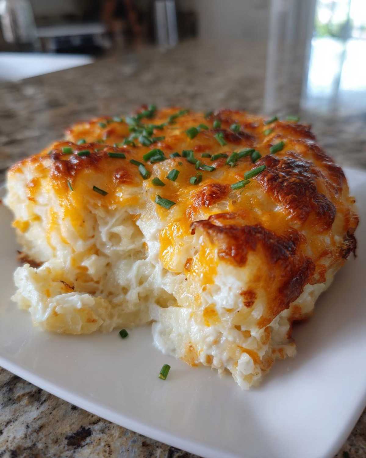 A close-up serving of cheesy potato casserole with a golden-brown crust, topped with fresh green chives.