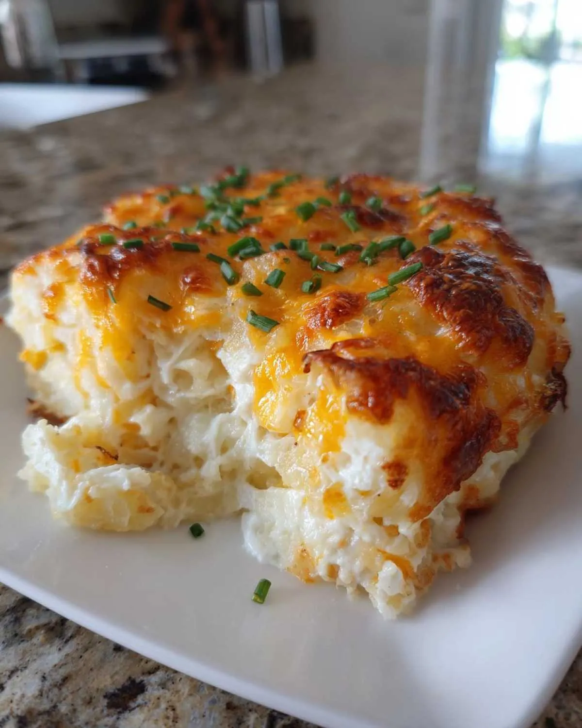 A close-up serving of cheesy potato casserole with a golden-brown crust, topped with fresh green chives.