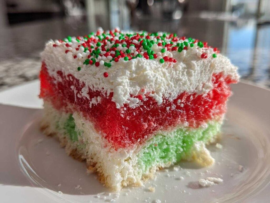 A close-up slice of christmas poke cake showing layers of white cake, red gelatin, green gelatin, and whipped topping with sprinkles.