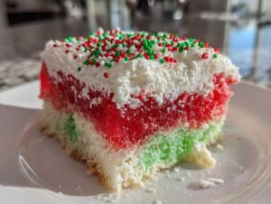 A close-up slice of christmas poke cake showing layers of white cake, red gelatin, green gelatin, and whipped topping with sprinkles.