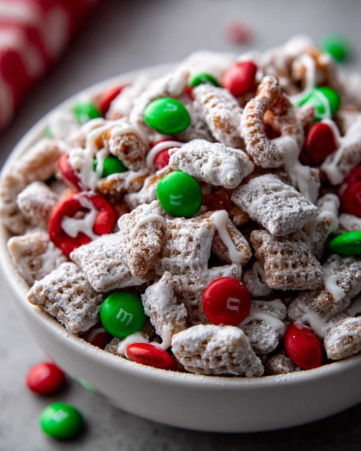 Close-up of a white bowl filled with Christmas puppy chow coated in powdered sugar and drizzled with white chocolate, mixed with red and green candies.