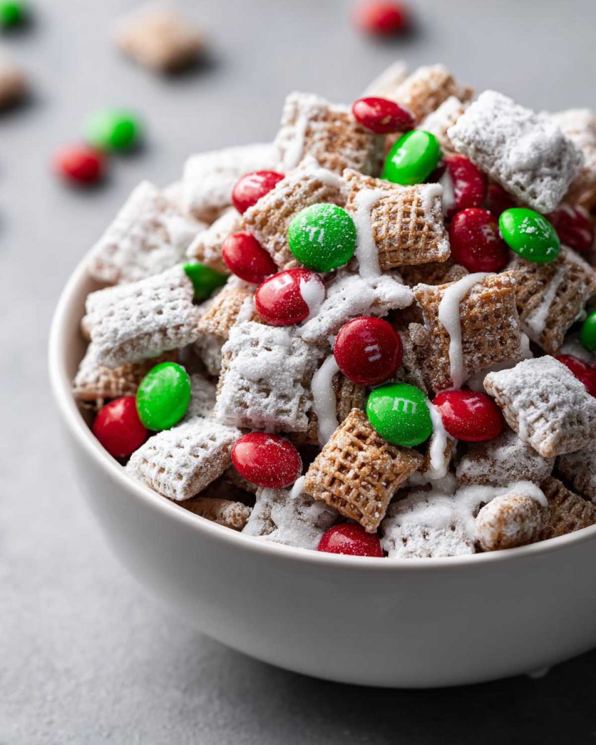 Close-up of a white bowl filled with powdered sugar coated Christmas Puppy Chow mixed with red and green M&Ms.