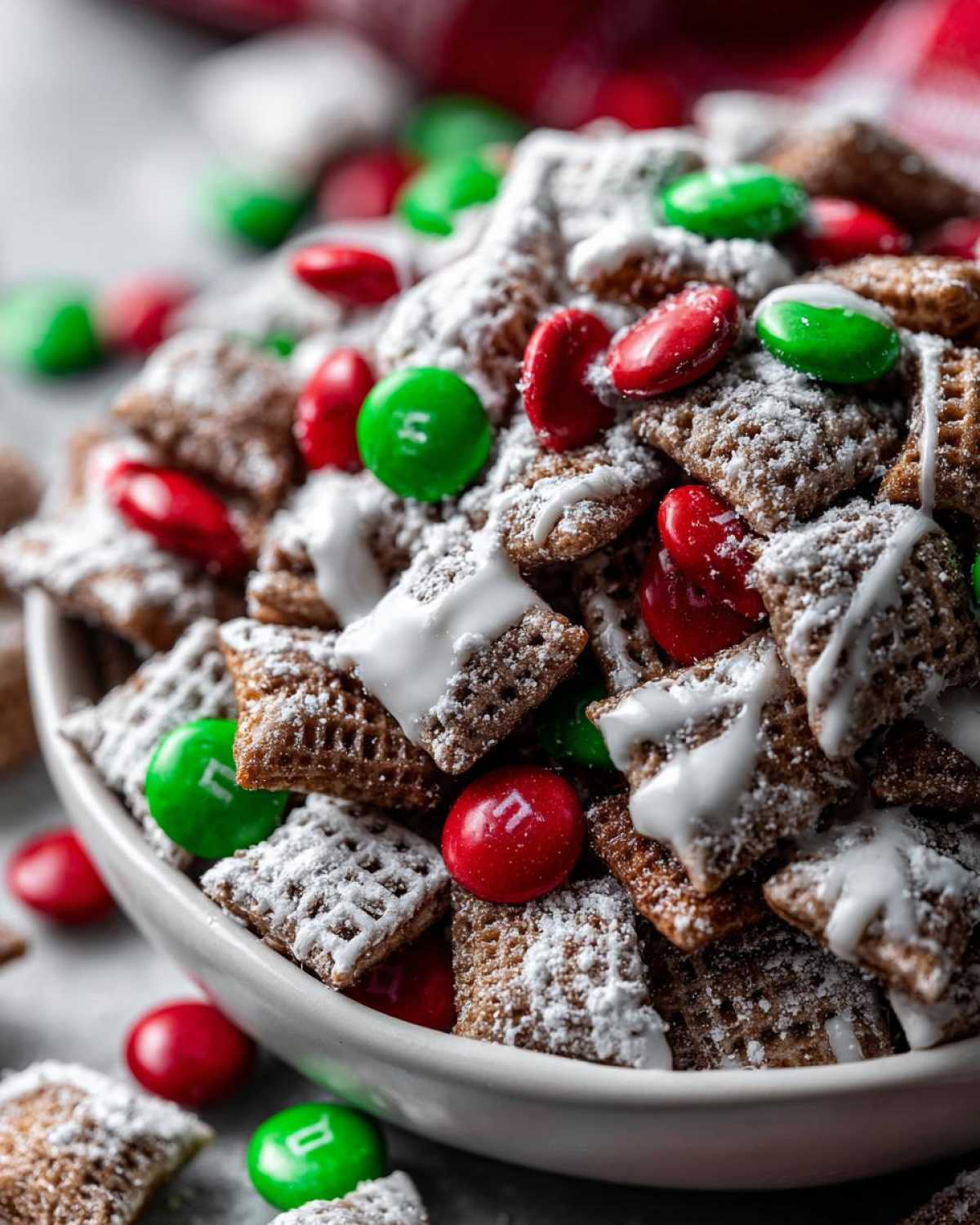 Close-up of a white bowl filled with Christmas puppy chow coated in powdered sugar and drizzled with white icing, mixed with red and green M&Ms.