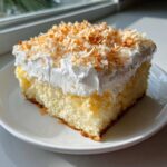 A generous slice of moist coconut poke cake topped with whipped cream and toasted coconut flakes.