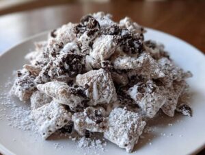 A mound of cookies and cream puppy chow coated in powdered sugar and dark cookie pieces, served on a white plate.