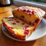 A loaf of moist cranberry orange bread, partially sliced, topped with a bright white orange glaze and visible cranberries.