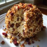 A creamy cranberry pecan cheese ball coated in nuts and dried cranberries, with a section scooped out, sitting on a white plate.