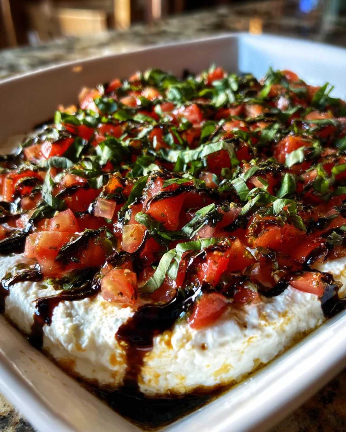 A close-up of a creamy bruschetta dip topped with diced tomatoes, fresh basil, and a rich balsamic glaze drizzle.