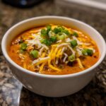 A close-up of a bowl of creamy buffalo chicken chili topped generously with shredded cheddar and Monterey Jack cheese and sliced green onions.