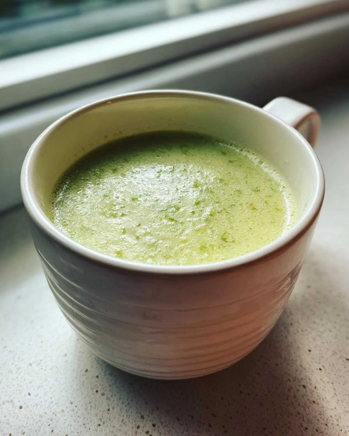 A close-up of creamy, light green celery soup served hot in a white, textured mug.