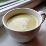A close-up of creamy, light green celery soup served hot in a white mug, resting on a windowsill.