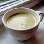 A close-up of creamy, light green celery soup served hot in a white mug, resting on a windowsill.