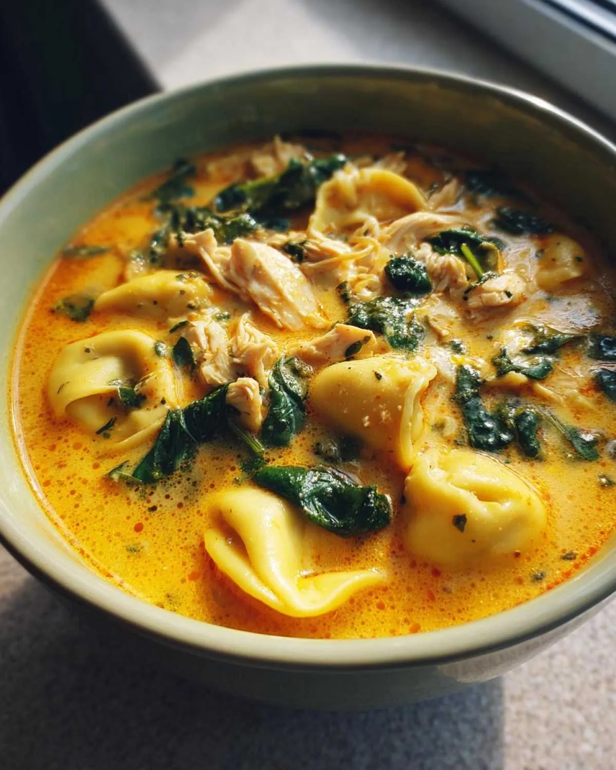 Close-up of a bowl of rich, orange-hued creamy chicken tortellini soup featuring tortellini pasta, shredded chicken, and wilted spinach.
