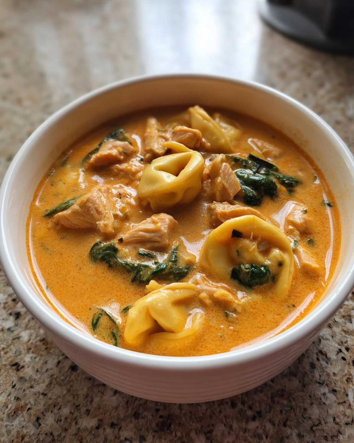 Close-up of a white bowl filled with creamy chicken tortellini soup, featuring pasta, chicken pieces, and spinach.