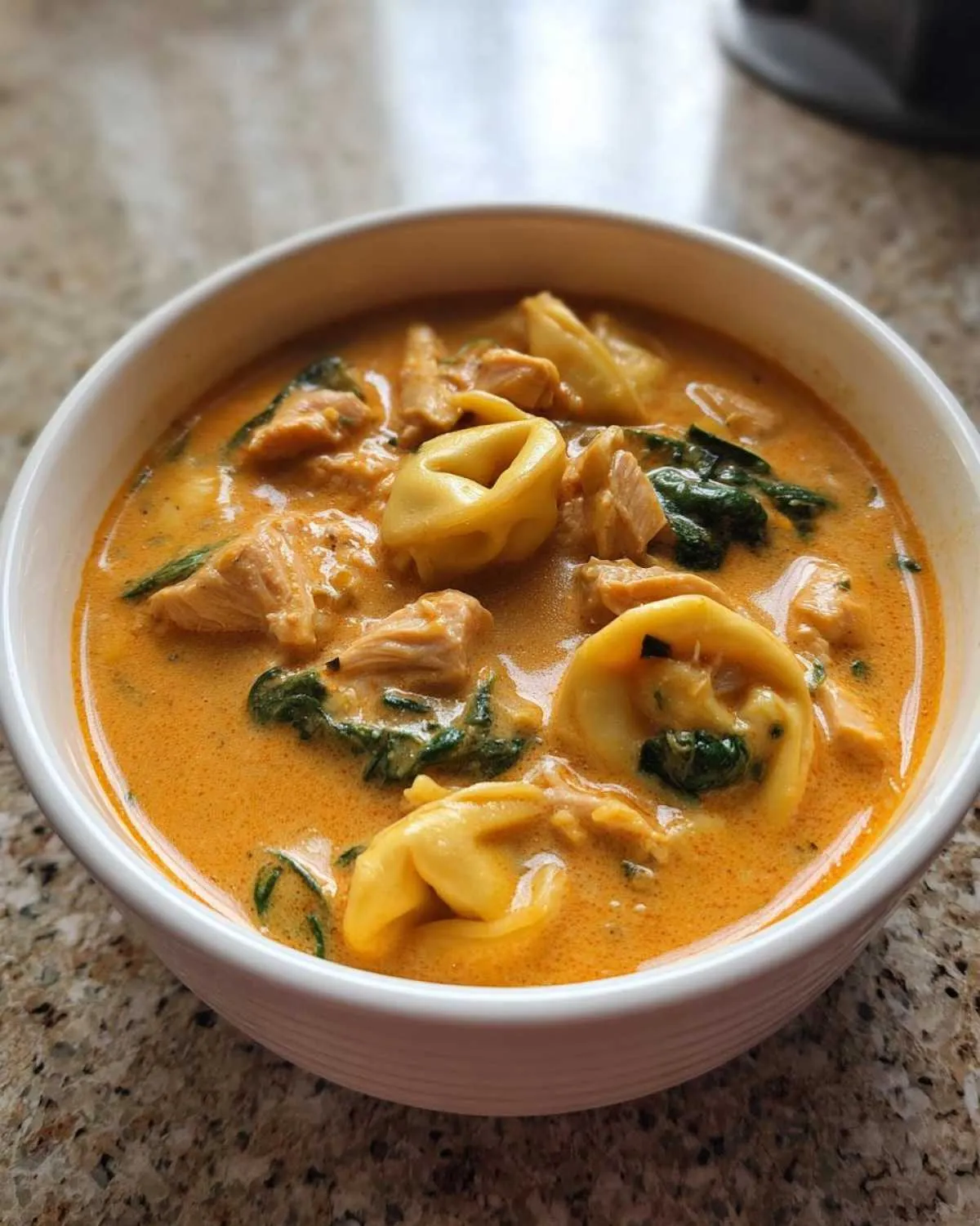 Close-up of a white bowl filled with creamy chicken tortellini soup, featuring pasta, chicken pieces, and spinach.