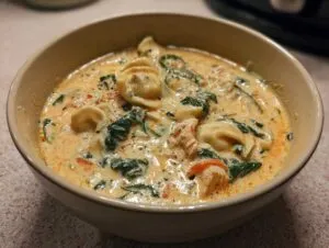Close-up of a beige bowl filled with creamy chicken tortellini soup, featuring tortellini, spinach, and chicken pieces.