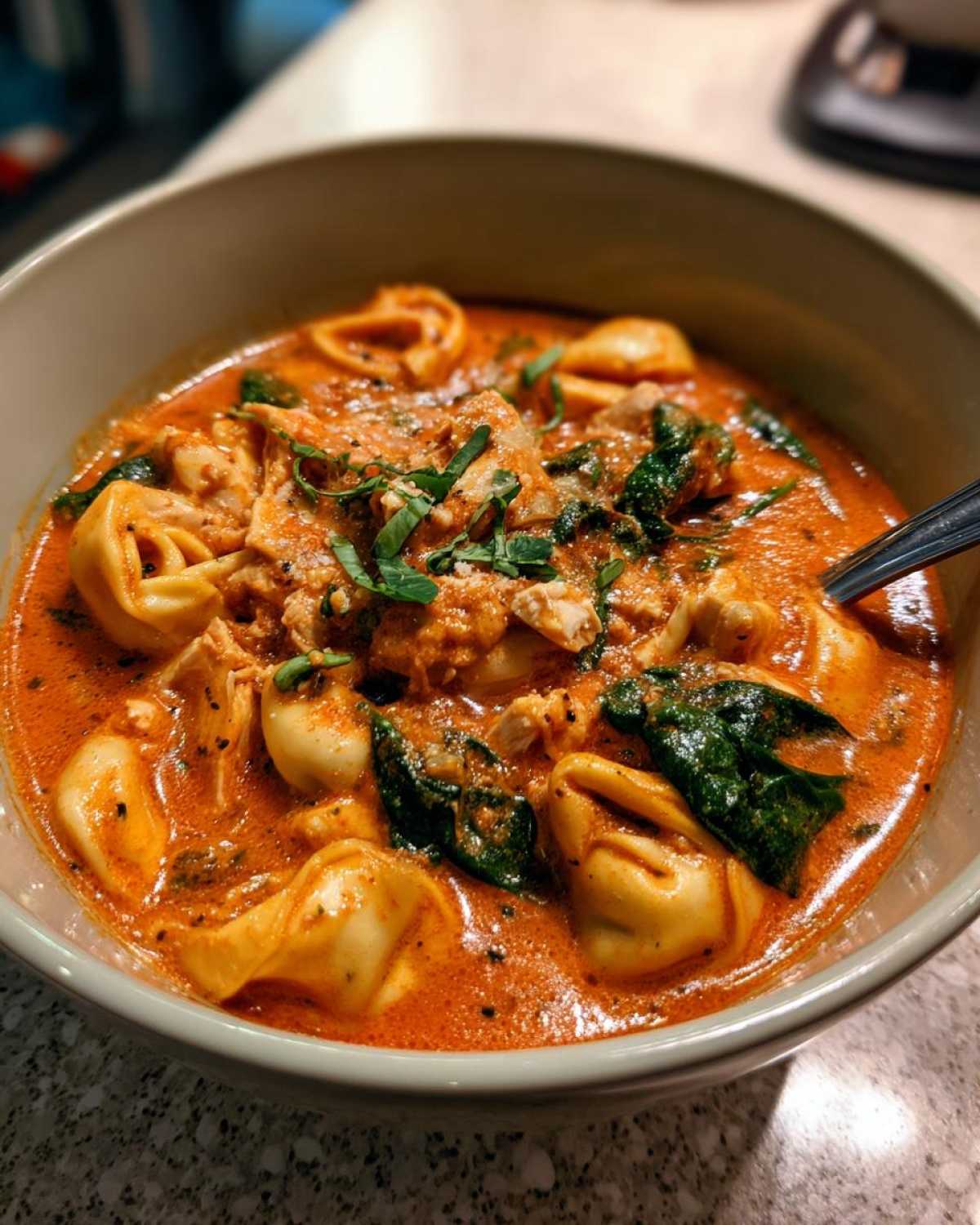 Close-up of a rich, orange-red bowl of creamy chicken tortellini soup garnished with fresh basil.