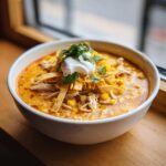 A close-up of a bowl of creamy chicken tortilla soup topped with shredded cheese, tortilla strips, sour cream, and cilantro.