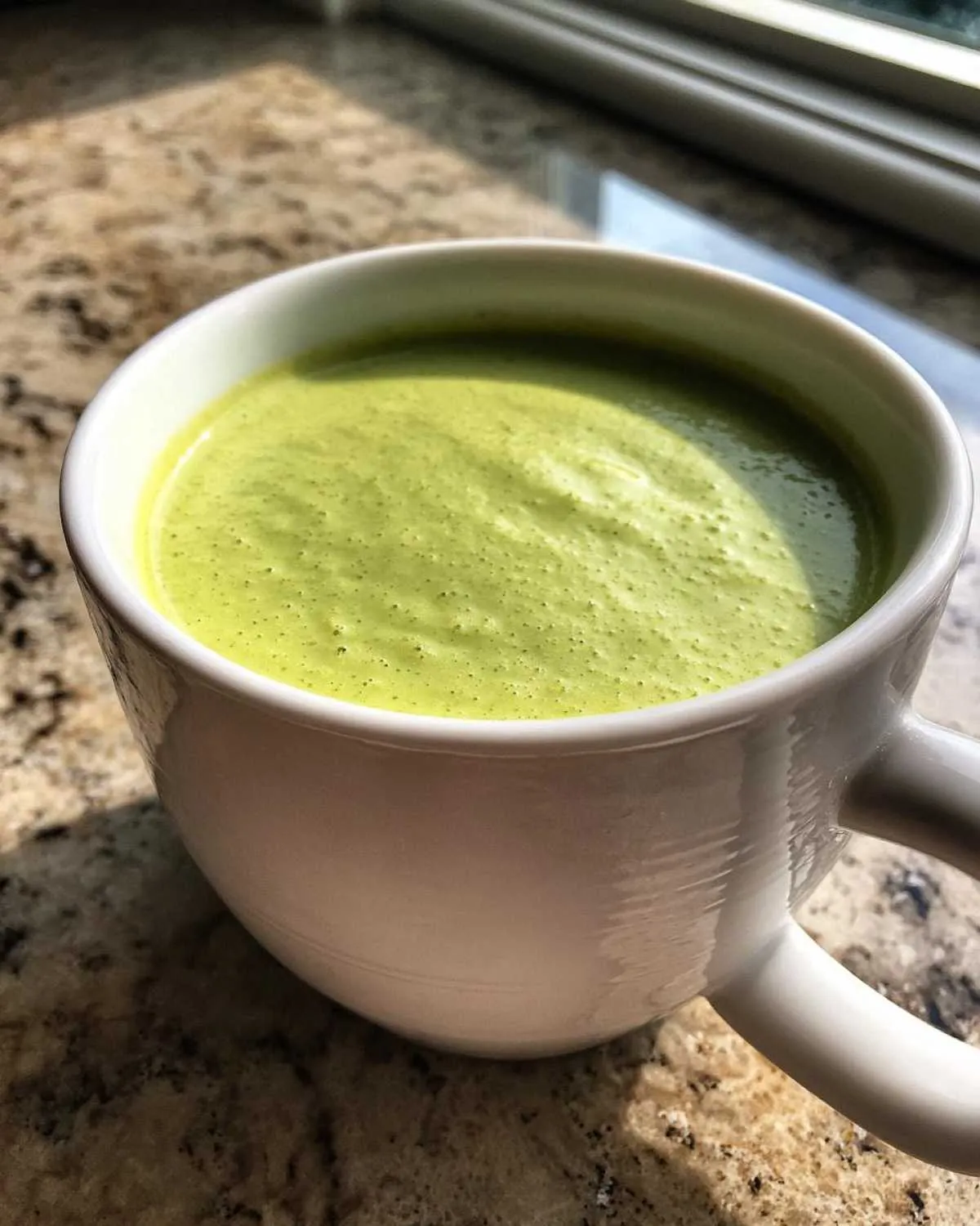 A close-up of creamy, bright green celery soup served hot in a white mug on a speckled countertop.