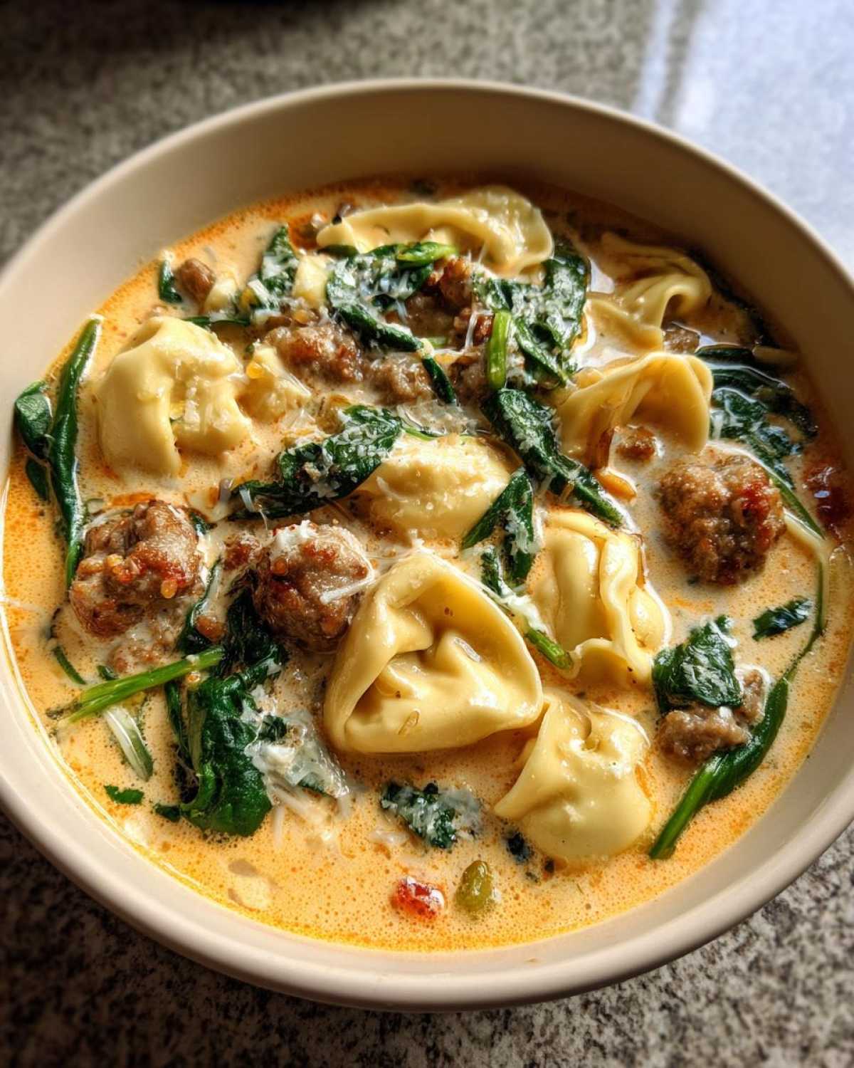 A close-up bowl of creamy italian sausage tortellini soup with spinach and grated cheese.