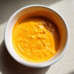A close-up overhead shot of thick, bright orange nacho cheese sauce filling a white bowl.