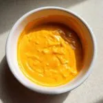 A close-up overhead shot of thick, bright orange nacho cheese sauce filling a white bowl.